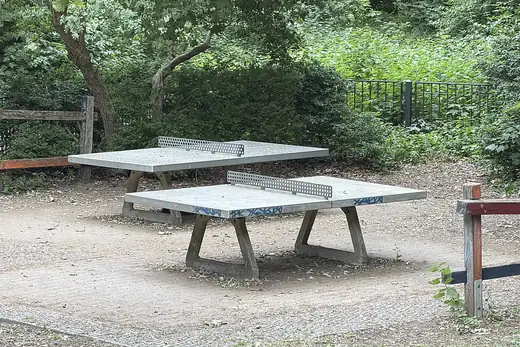 Zwei Betontische mit Metallnetzen auf einem Kiesplatz unter Bäumen